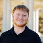 Elliott smiles at the camera, wearing a black collared shortsleeve shirt and green leaf earrings